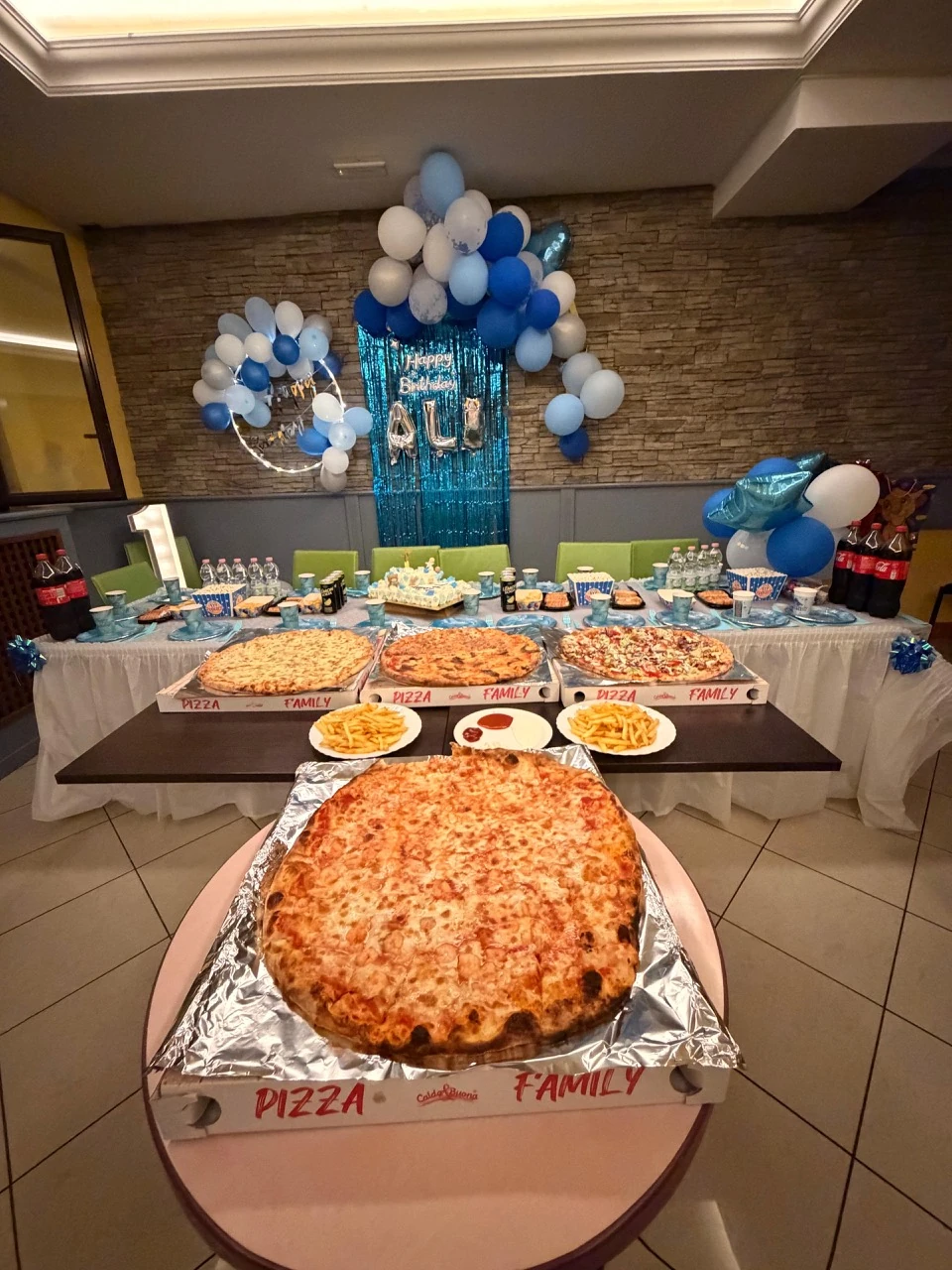 Une table d’anniversaire chez Mimmo, avec des pizzas familiales et des ballons bleus et blancs.